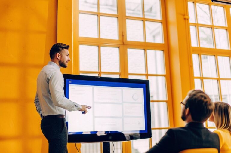 Νέος επιχειρηματίας της TWO DOTS παρουσιάζει την ιστοσελίδα σε συναδέλφους σε μοντέρνο γραφείο, δείχνοντας αξιοπιστία.