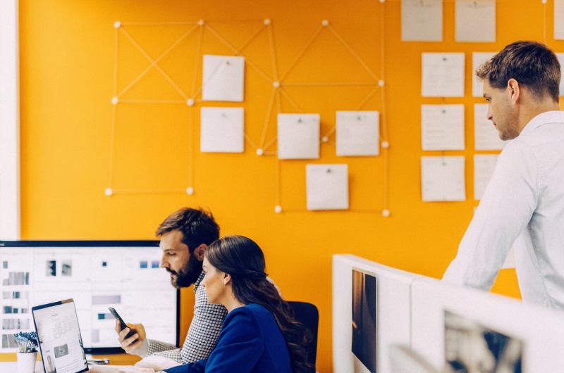 Εύστοχα ντυμένοι επιχειρηματίες και επιχειρηματίες κάθε φύλου δουλεύουν μαζί σε γραφείο με σύγχρονη διακόσμηση, κοιτώντας οθόνες υπολογιστών και κινητά που εμφανίζουν γραφήματα και δημοσιεύσεις του Instagram