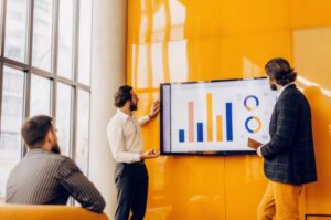 Σύγχρονοι επιχειρηματίες σε ένα γραφείο, κοιτάζουν μια μεγάλη οθόνη με πολύχρωμα διαγράμματα, γραφήματα και στατιστικά, συζητώντας στρατηγικές marketing και KPIs