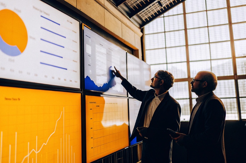 Ρεαλιστική φωτογραφία επιχειρηματιών σε σύγχρονο γραφείο με μεγάλες οθόνες που προβάλλουν γραφήματα, analytics και στατιστικά στοιχεία για το μερίδιο φωνής τεχνητής νοημοσύνης, οι επιχειρηματίες συζητούν μεταξύ τους και δείχνουν σε δεδομένα AI, περιβάλλον υψηλής τεχνολογίας, επαγγελματική ατμόσφαιρα, φυσικός φωτισμός