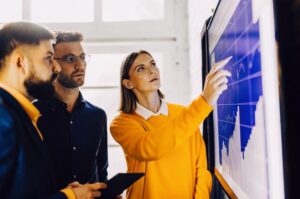 Ομάδα επιχειρηματιών και επαγγελματιών σε σύγχρονο γραφείο παρακολουθούν οθόνη με γραφήματα και αναλύσεις δεδομένων αλληλεπιδρούν με προηγμένα ψηφιακά εργαλεία