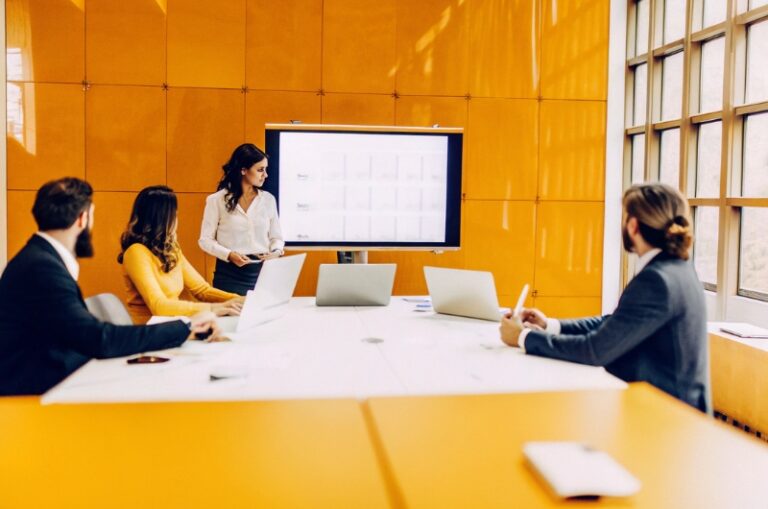 Επαγγελματίες και επιχειρηματίες σε ένα σύγχρονο γραφείο συνεργάζονται γύρω από τραπέζι γεμάτο φορητούς υπολογιστές και σημειώσεις, ενώ παρακολουθούν παρουσιάσεις σε οθόνη