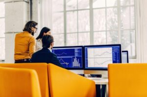 Επαγγελματίες και επιχειρηματίες σε ένα σύγχρονο γραφείο συνεργάζονται μπροστά σε οθόνες υπολογιστών όπου εμφανίζονται διαγράμματα, αναλύσεις δεδομένων και ψηφιακά γραφήματα